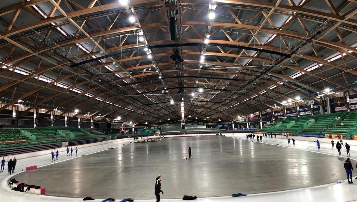 Indoor skating rink with skaters on the ice. Overhead lighting and wooden roof structures are visible above the rink.