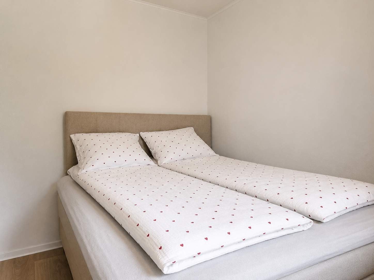 Two twin beds with white heart-pattern bedding and pillows sit against a beige headboard in a plain room.