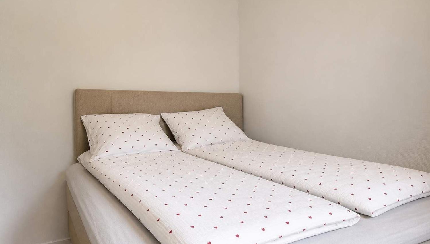 Two twin beds with white heart-pattern bedding and pillows sit against a beige headboard in a plain room.
