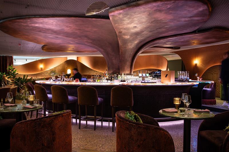 En elegant bar med organiske, bølgende takformer, dempet belysning og luksuriøse sitteplasser skaper en intim og moderne atmosfære.