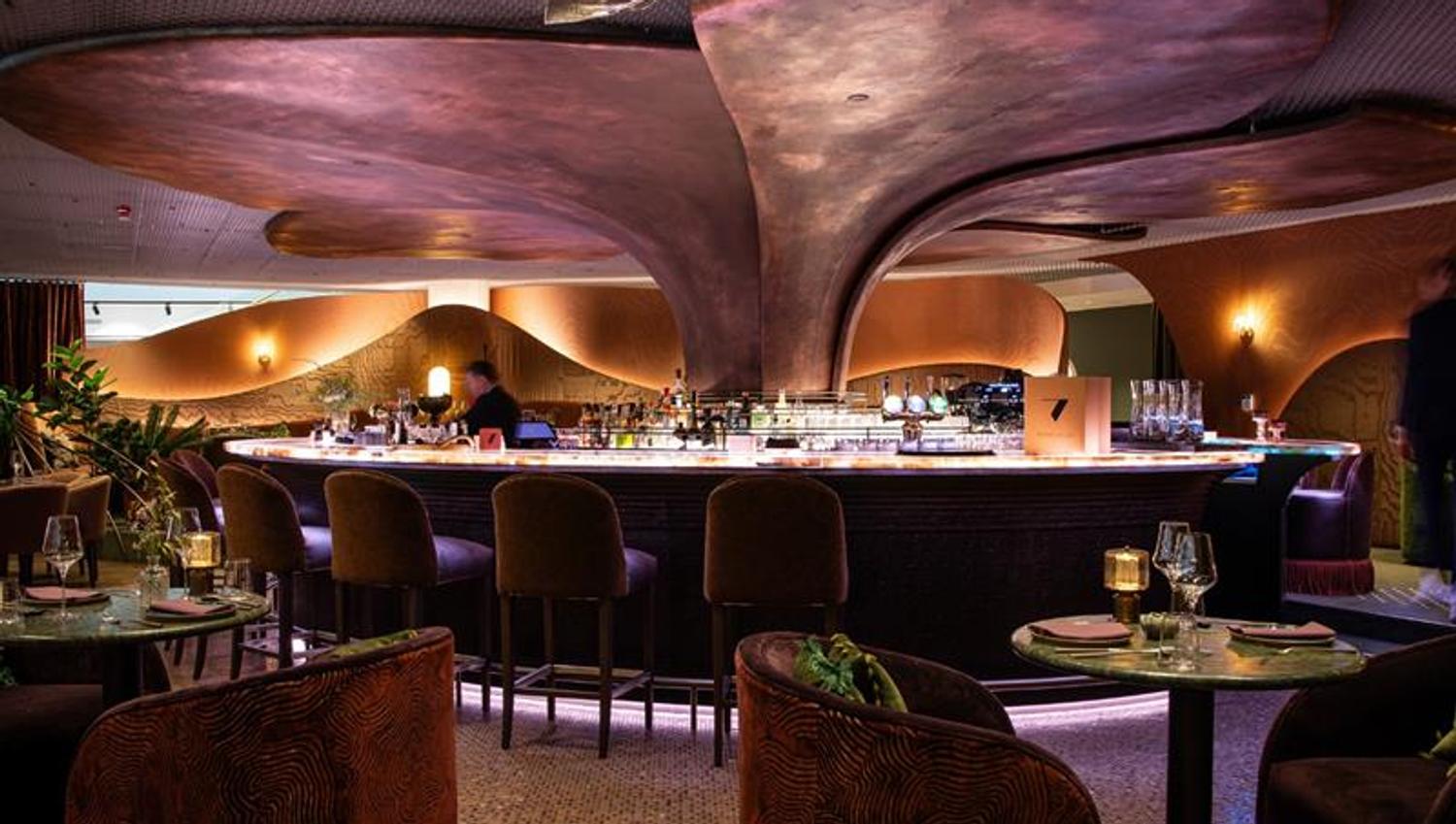 En elegant bar med organiske, bølgende takformer, dempet belysning og luksuriøse sitteplasser skaper en intim og moderne atmosfære.