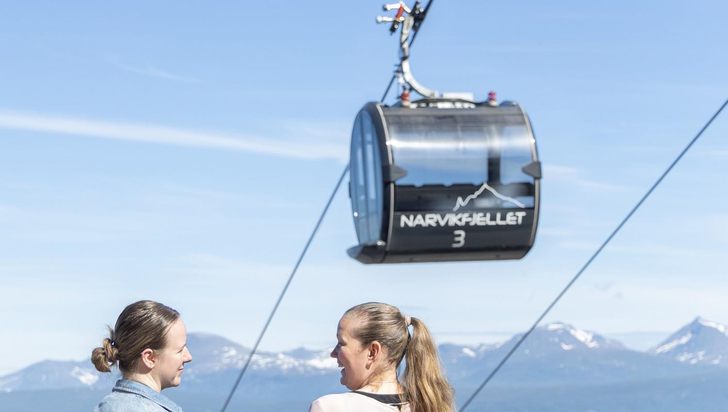 Two women talk by a railing with drinks while a gondola cabin hangs overhead against blue sky and snowy mountains.