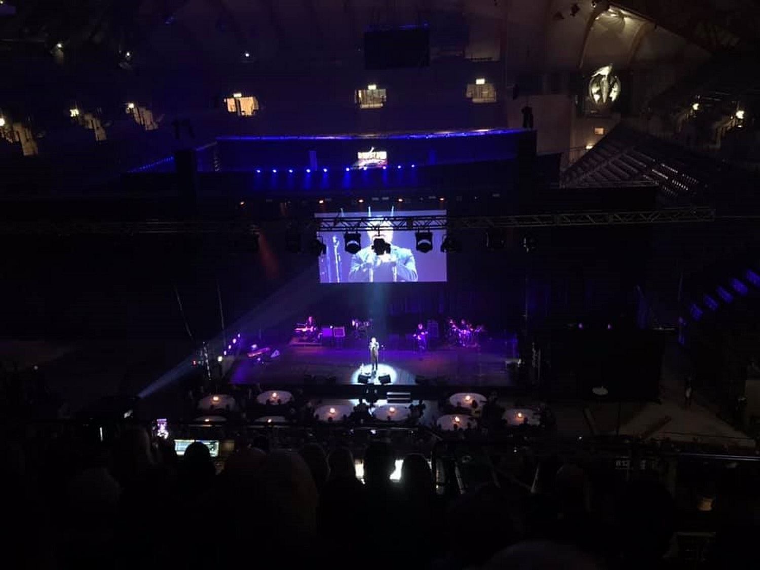 Mørk konsertsal med scene i lilla lys, en artist i spotlight, band på siden og stor skjerm over scenen..