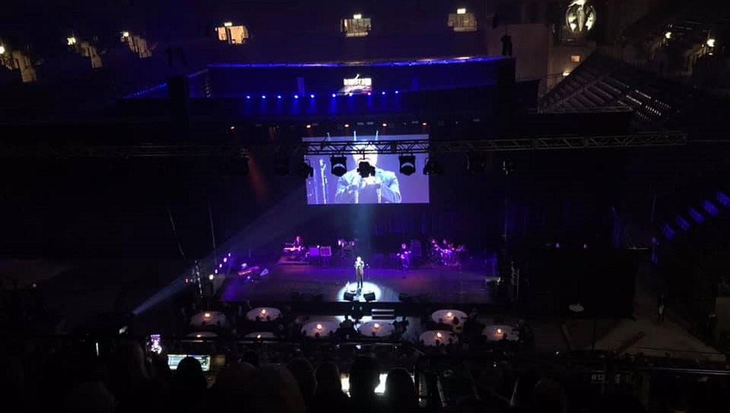 Mørk konsertsal med scene i lilla lys, en artist i spotlight, band på siden og stor skjerm over scenen..