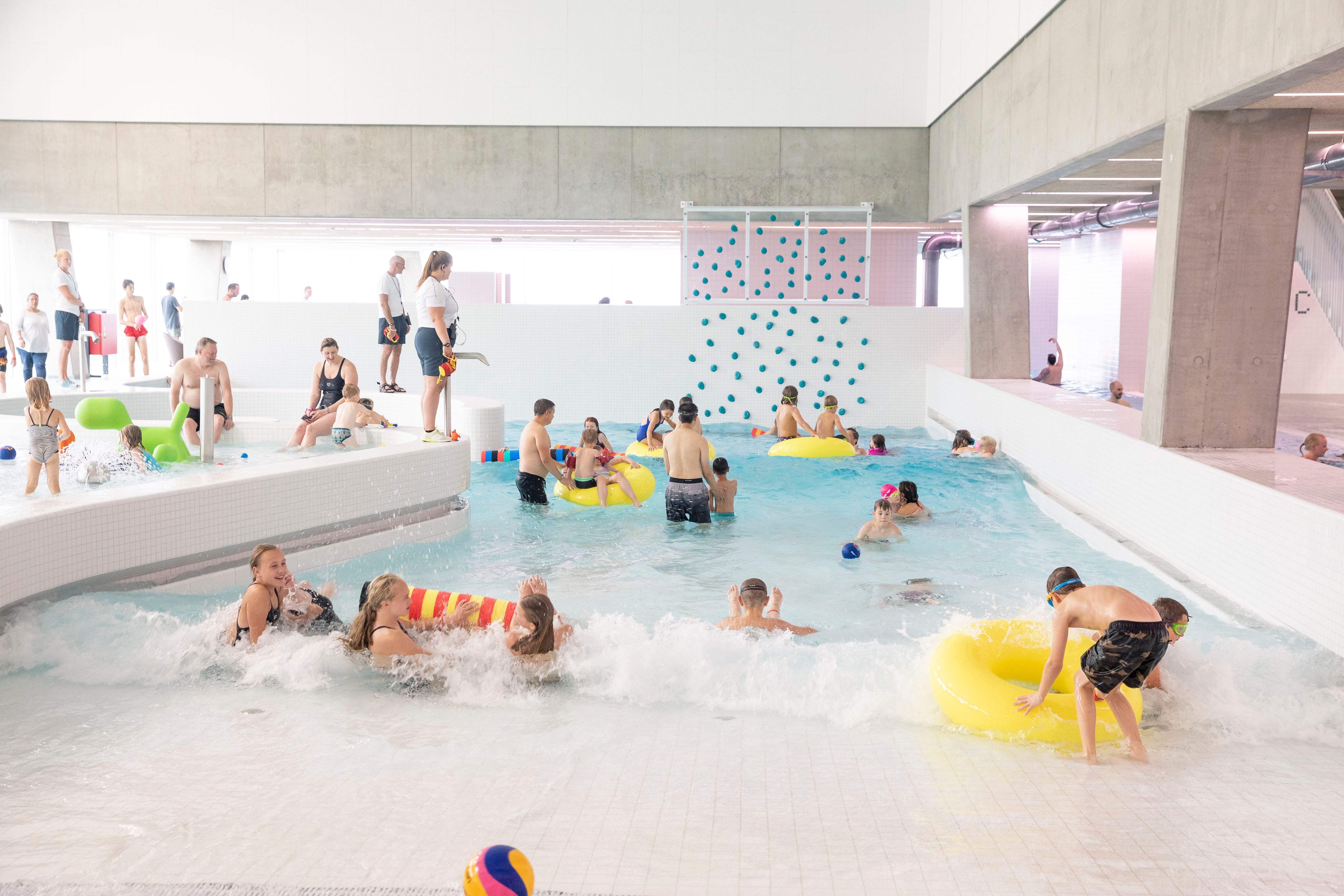 People of all ages enjoying a bright indoor swimming pool with a climbing wall