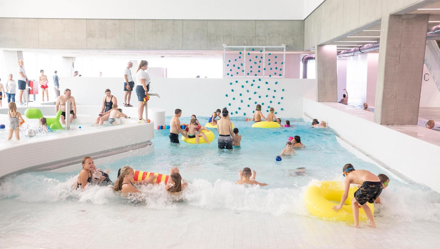 People of all ages enjoying a bright indoor swimming pool with a climbing wall