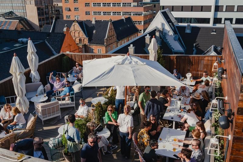 The rooftop terrace features seating areas, parasols and greenery, with views across the surrounding rooftops. The space is designed for service and s