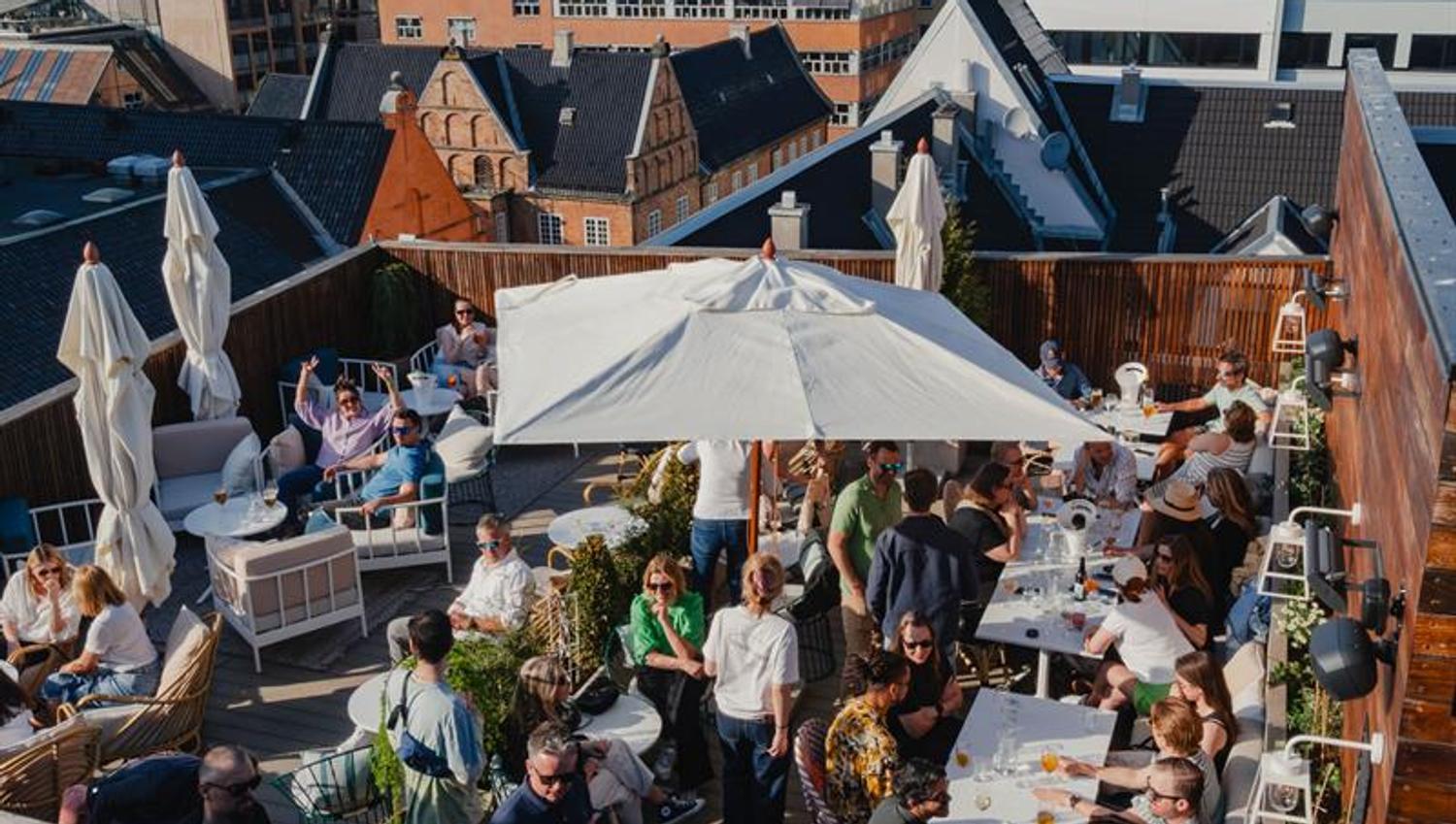The rooftop terrace features seating areas, parasols and greenery, with views across the surrounding rooftops. The space is designed for service and s