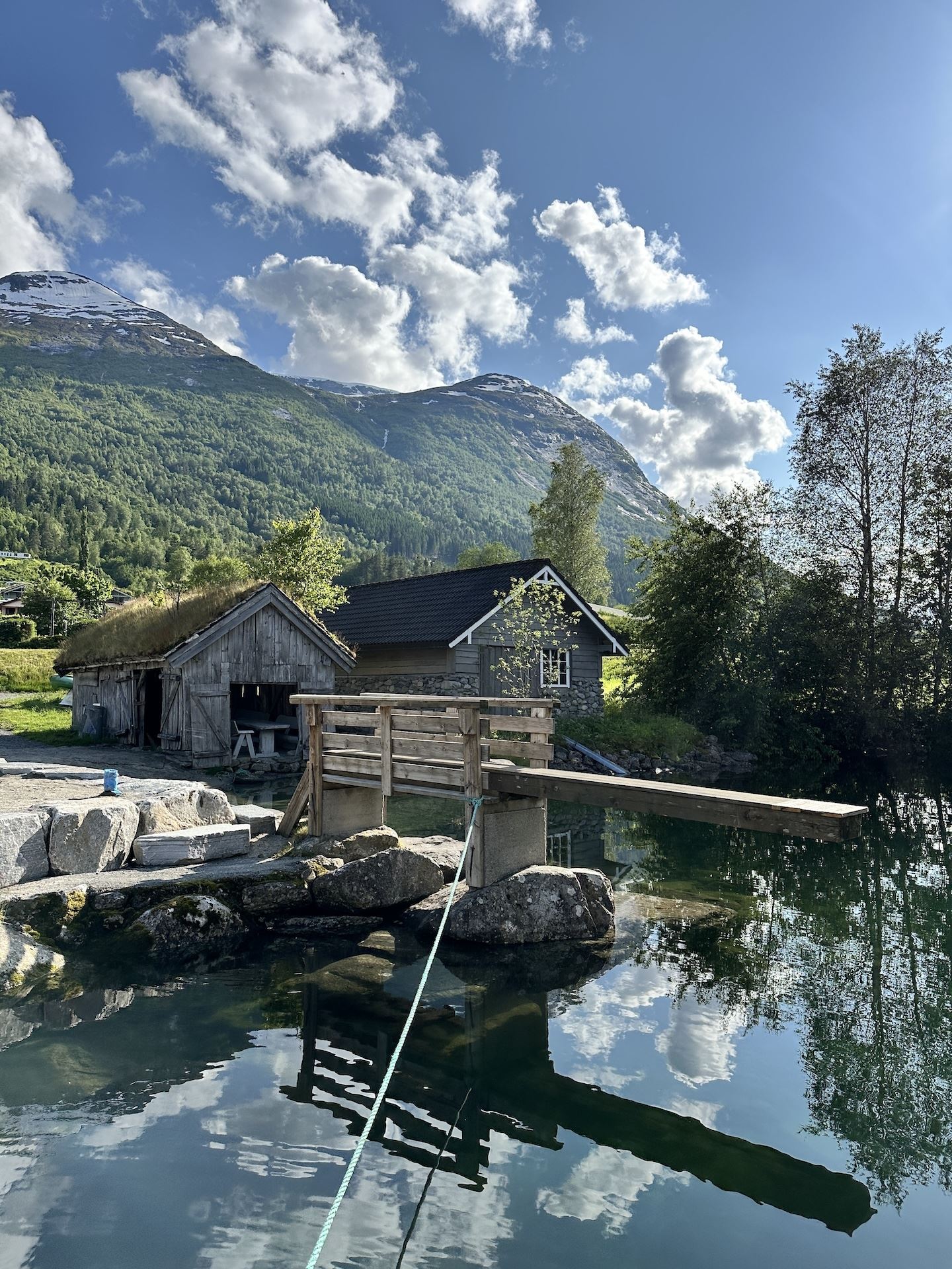 Rustic wooden cabins and a small footbridge by a clear pond, with green mountains and scattered clouds overhead..