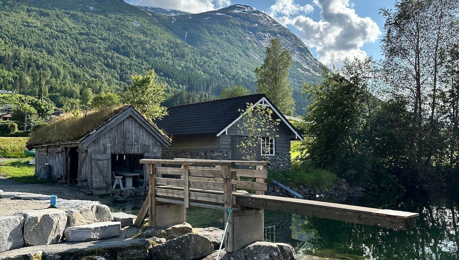 Rustic wooden cabins and a small footbridge by a clear pond, with green mountains and scattered clouds overhead..