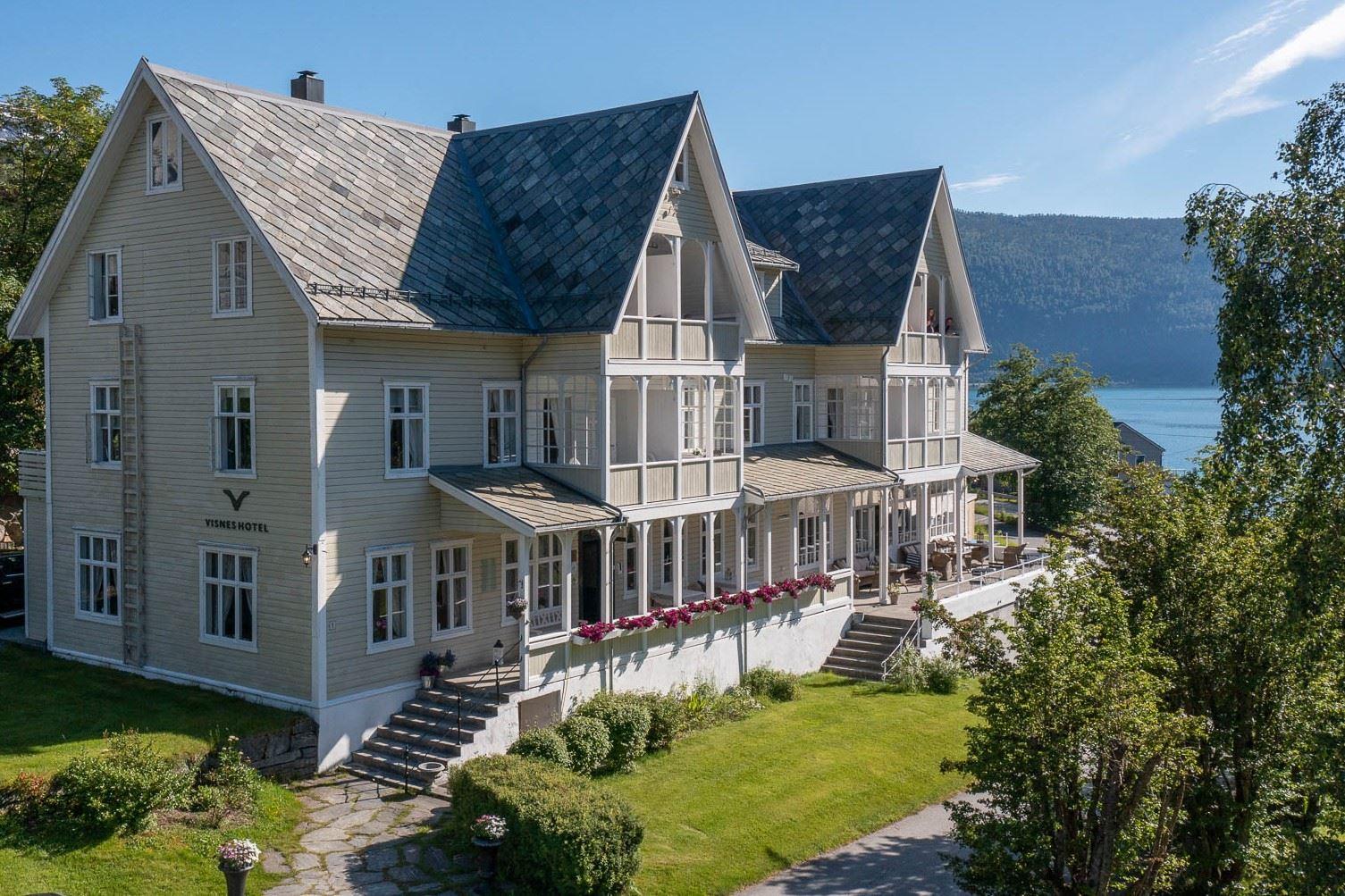 Large light-gray wooden Visnes Hotel Stryn with steep gabled roofs, balconies, and garden lawn beside a lake and hills..