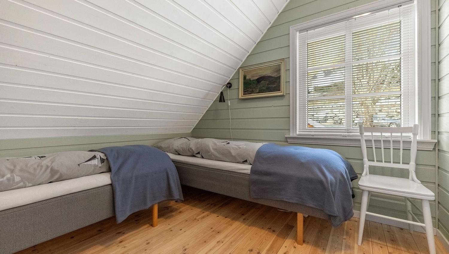 Small attic bedroom with two single beds, blue blankets, wood floor, window blinds, and a white chair at the Seahouse at Hornelen Panorama in Bremanger..
