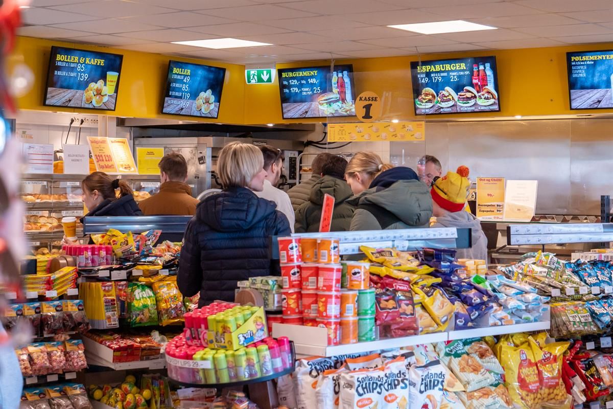 A busy convenience store interior where customers are shopping in front of shelves and counters stocked with snacks, drinks, and fast food items.