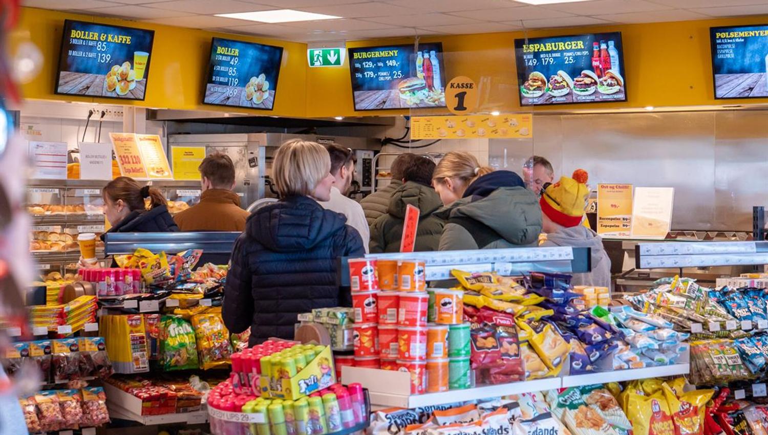 A busy convenience store interior where customers are shopping in front of shelves and counters stocked with snacks, drinks, and fast food items.
