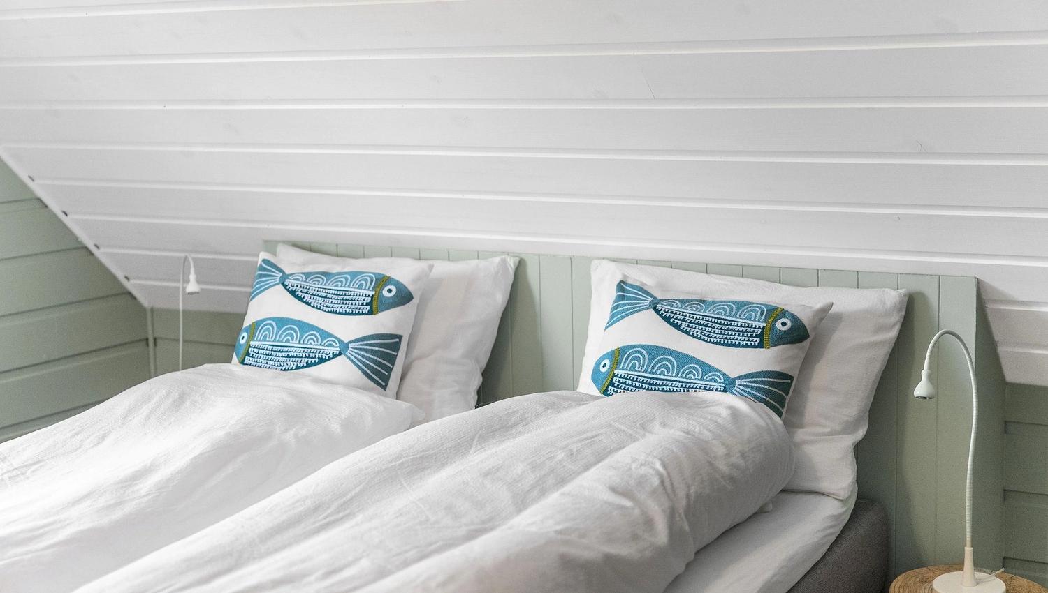 Two neatly made twin beds with white bedding and blue fish-pattern pillows under a slanted white wood ceiling at the Seahouse at Hornelen Panorama in Bremanger..