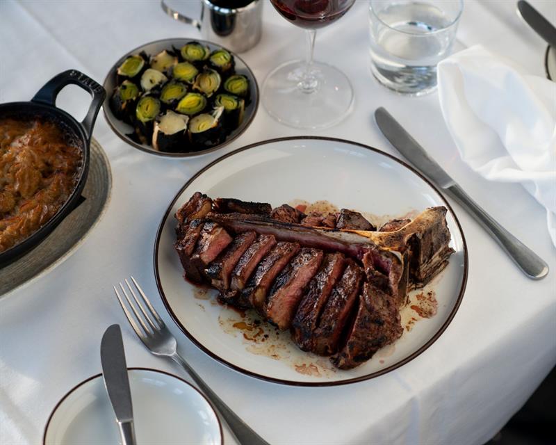 A grilled côte de boeuf served on a white plate with side dishes and wine glasses on a set table.
