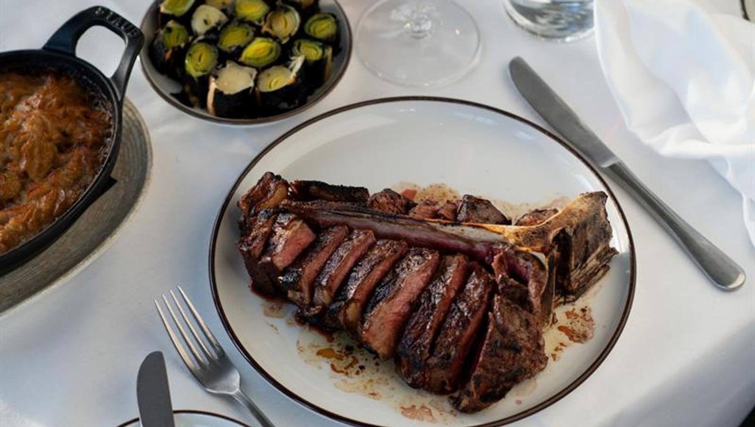 A grilled côte de boeuf served on a white plate with side dishes and wine glasses on a set table.