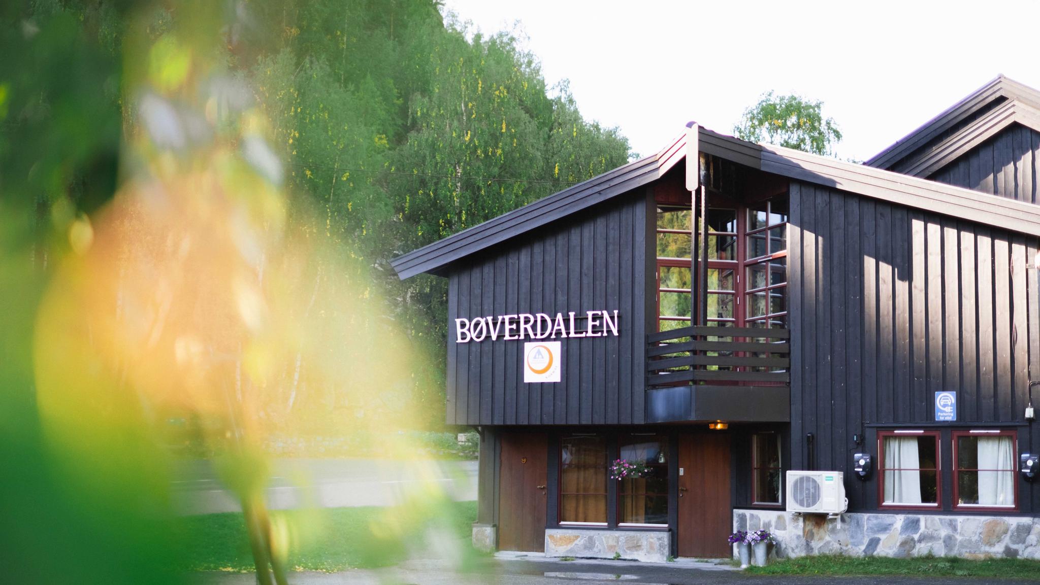 Black wooden building with the sign “BØVERDALEN,” large windows, and green trees behind in daylight..