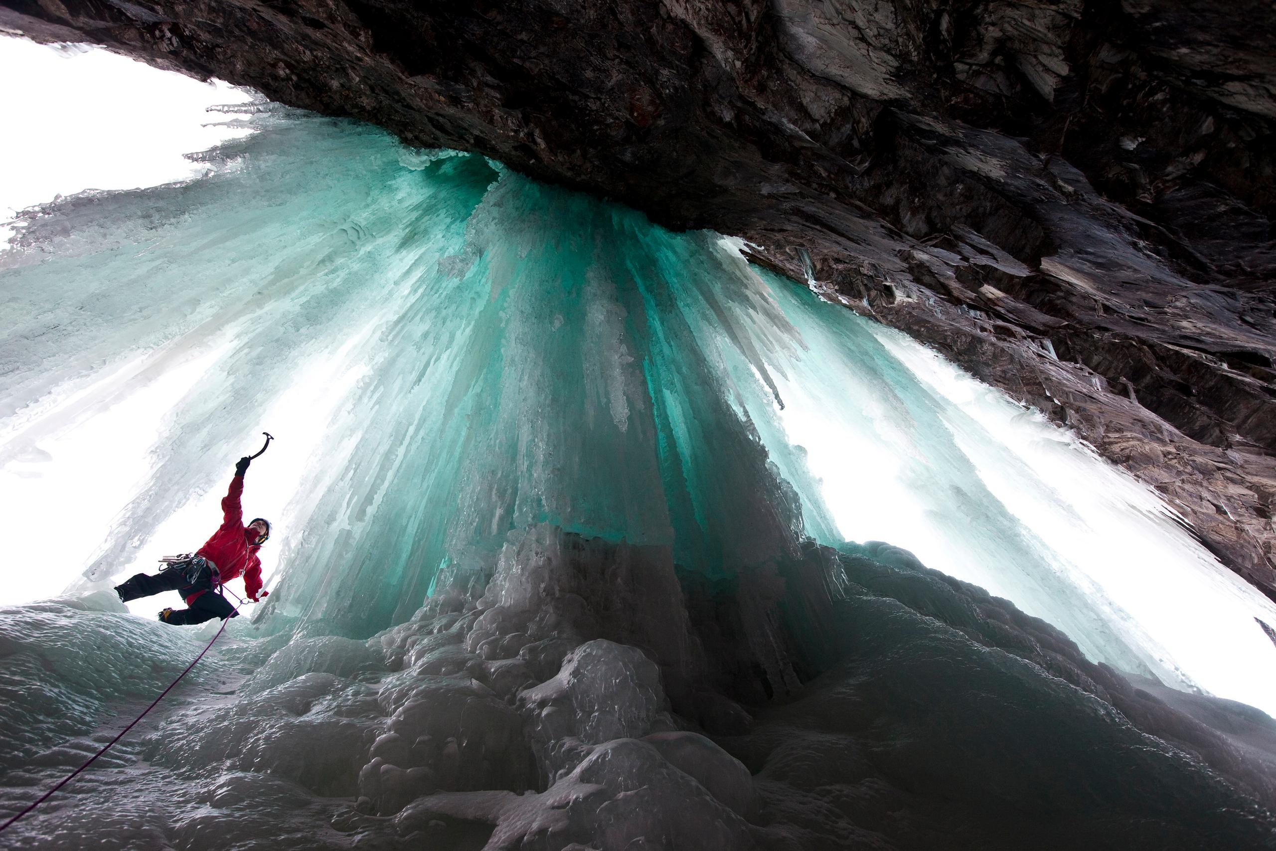 An iceclimber in a red jacket seen from below climbing the turquoise colored ice.