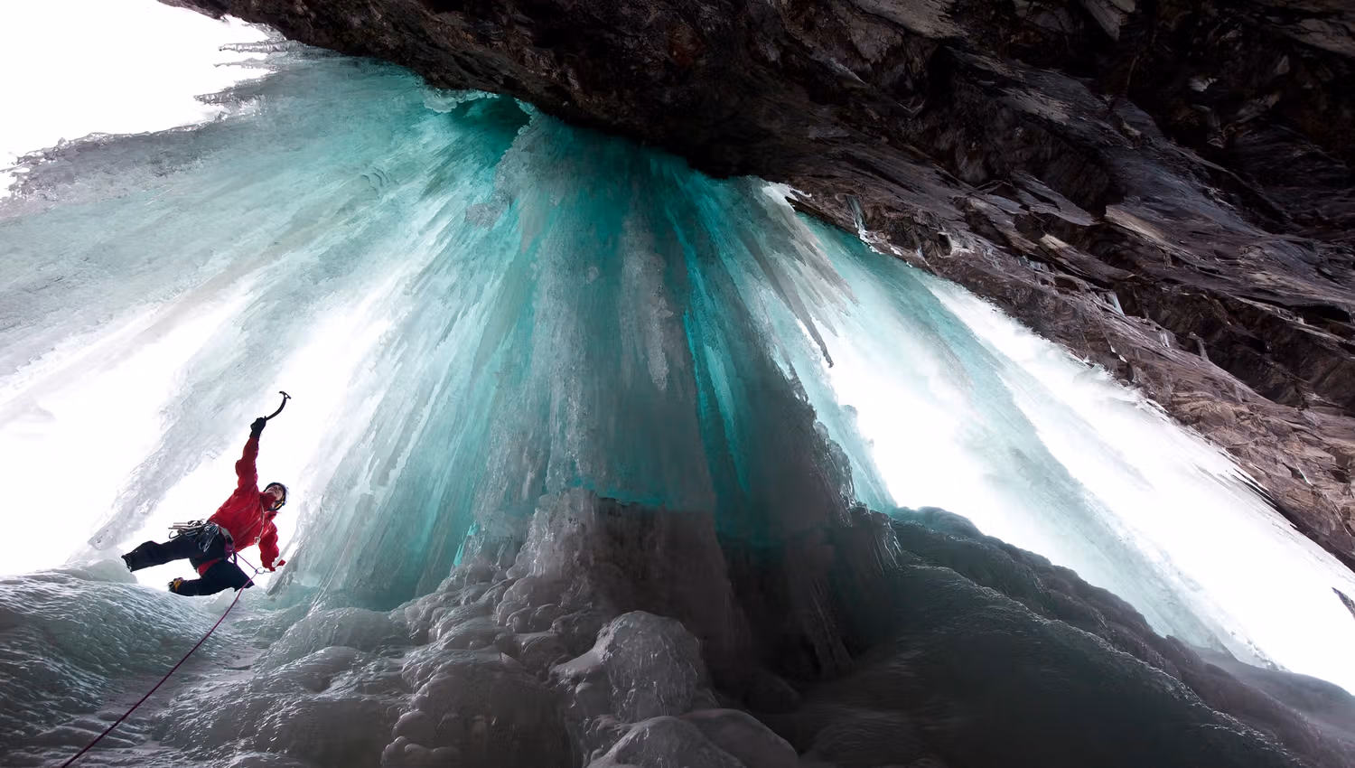 An iceclimber in a red jacket seen from below climbing the turquoise colored ice.