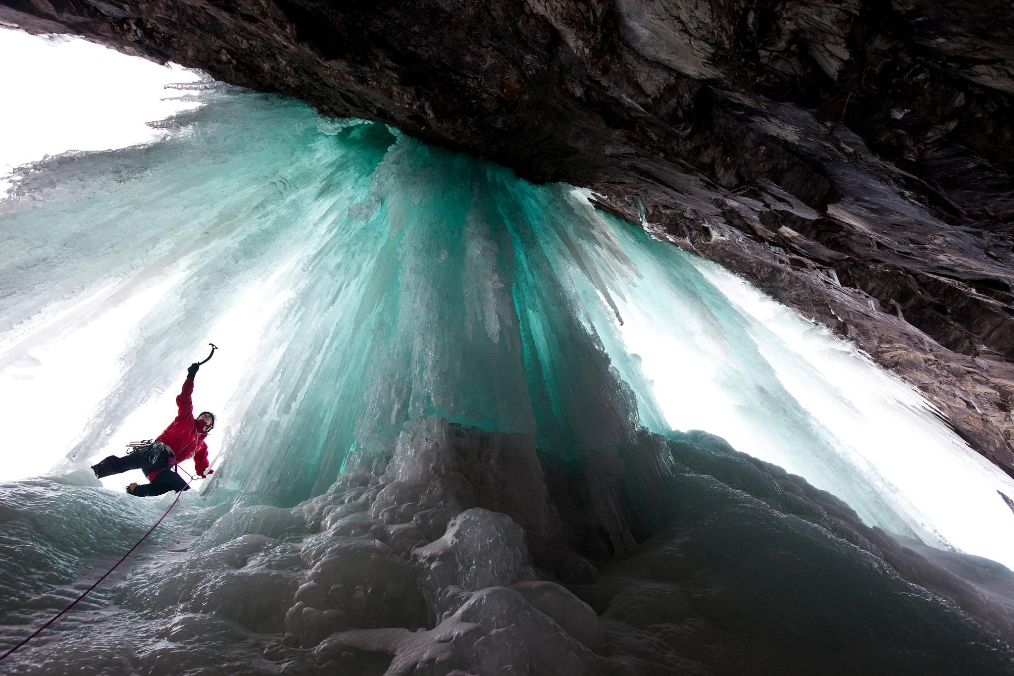 An iceclimber in a red jacket seen from below climbing the turquoise colored ice.