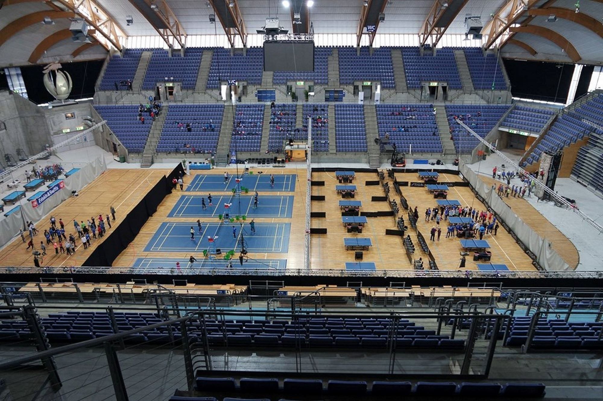 Stor innendørs idrettsarena med tribuner, badmintonbaner og bordtennisbord, samt grupper av deltakere på gulvet..