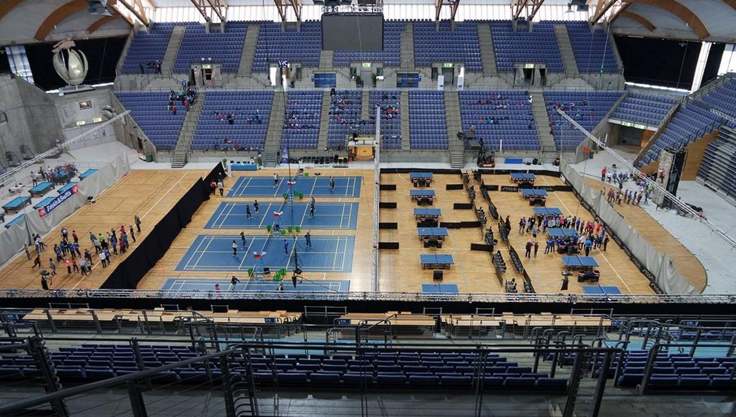Stor innendørs idrettsarena med tribuner, badmintonbaner og bordtennisbord, samt grupper av deltakere på gulvet..