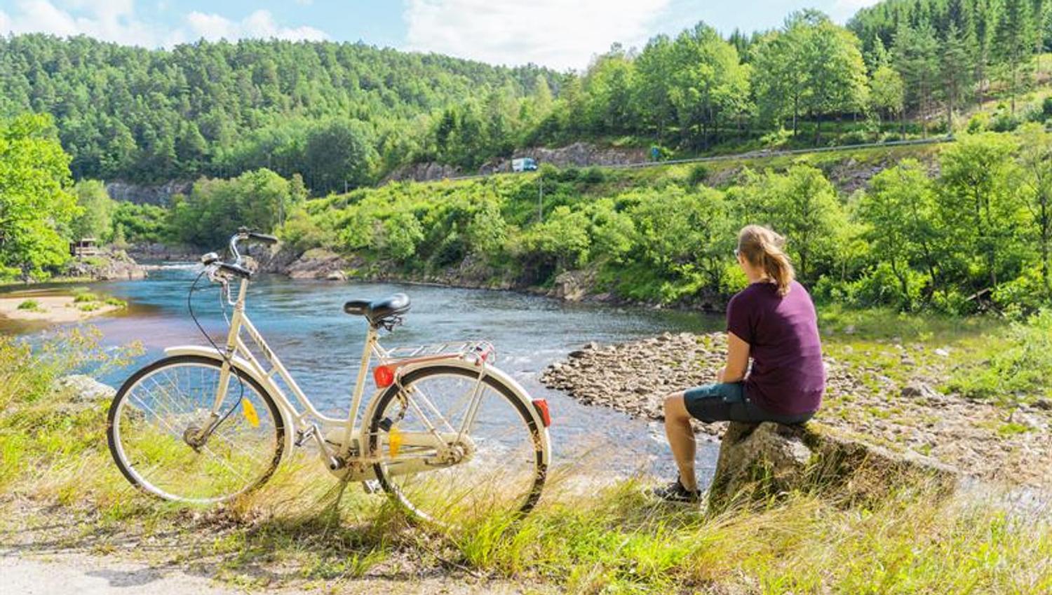 En dame sittende på en stein og ser mot Mandalselva, hvit sykkel vedsiden av henne.