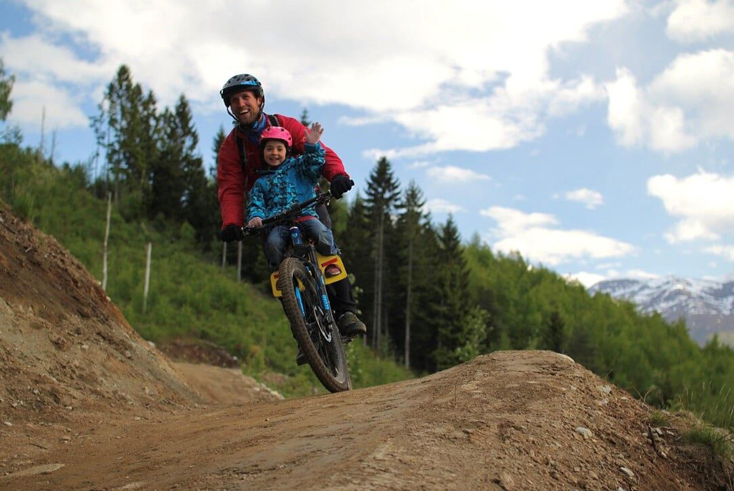 Adult and child wearing helmets ride a mountain bike over a dirt jump, with pine trees and cloudy sky behind.