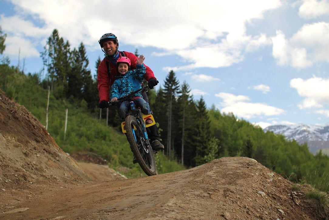 Adult and child wearing helmets ride a mountain bike over a dirt jump, with pine trees and cloudy sky behind.