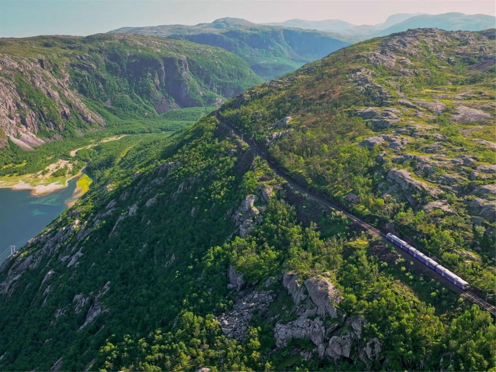 A passenger train travels along a winding track on a green, rocky mountainside above a valley and water.