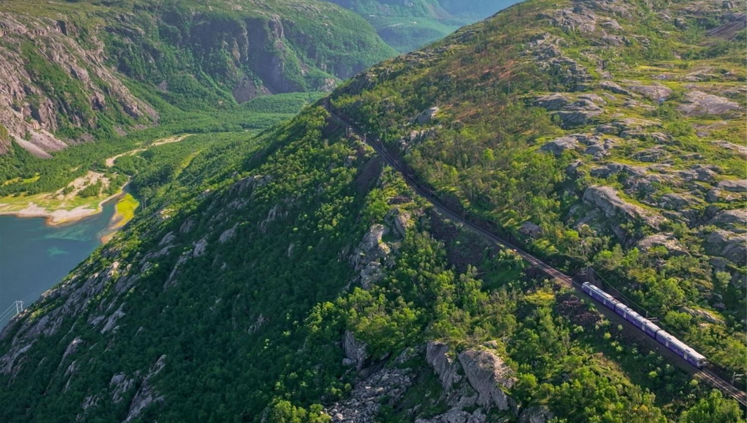 A passenger train travels along a winding track on a green, rocky mountainside above a valley and water.