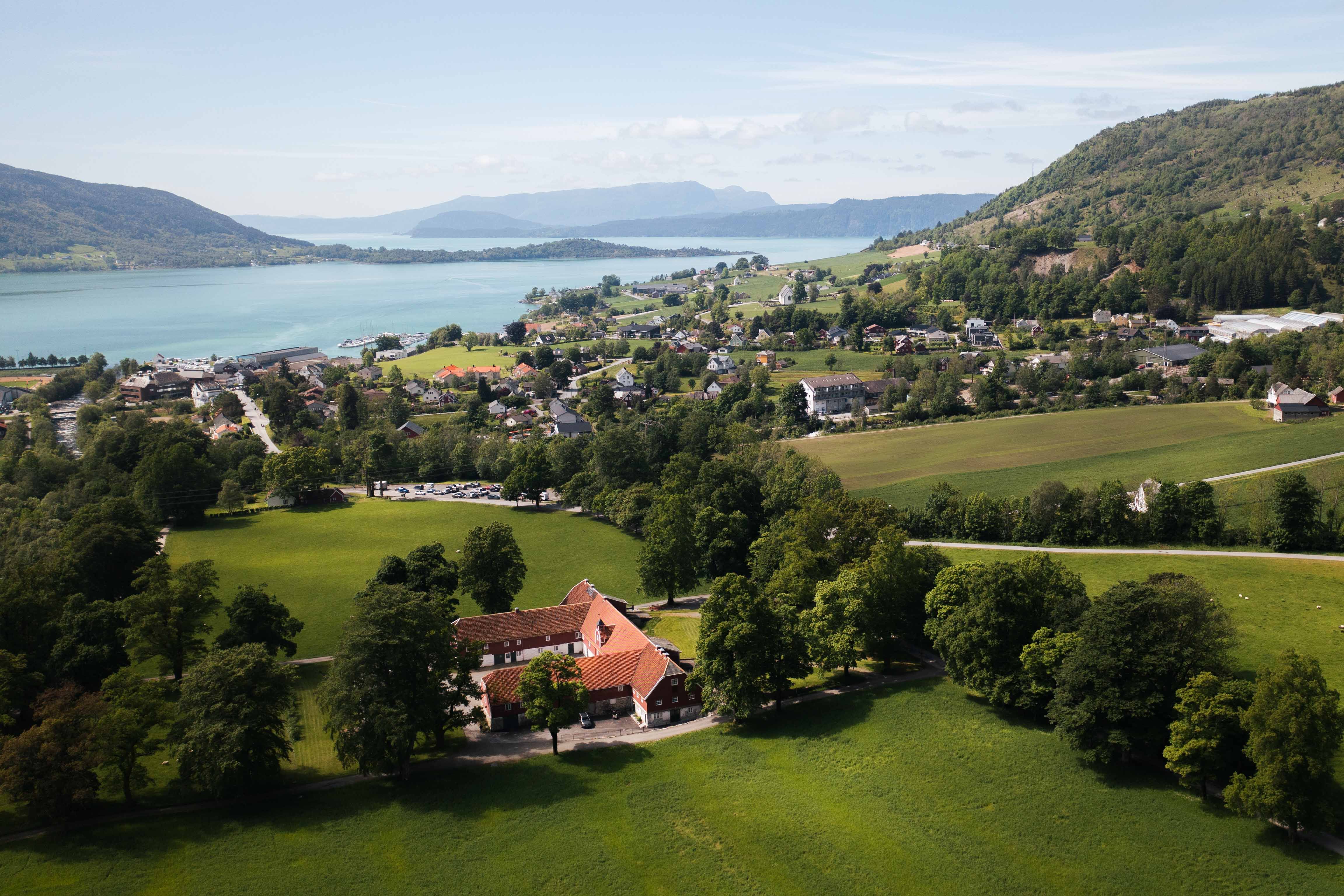 Dronebilete av Baroniet Rosendal Avlsgård og Fruehus – Storslått utsikt over det historiske gardsanlegget, omgitt av grøne marker og Hardangerfjorden i bakgrunnen.
