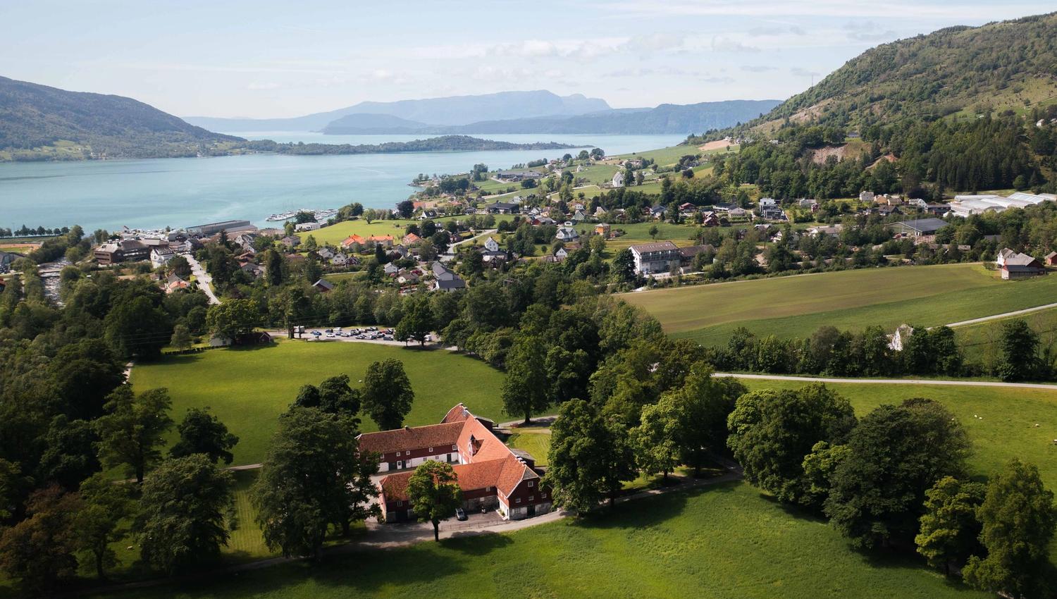 Dronebilete av Baroniet Rosendal Avlsgård og Fruehus – Storslått utsikt over det historiske gardsanlegget, omgitt av grøne marker og Hardangerfjorden i bakgrunnen.