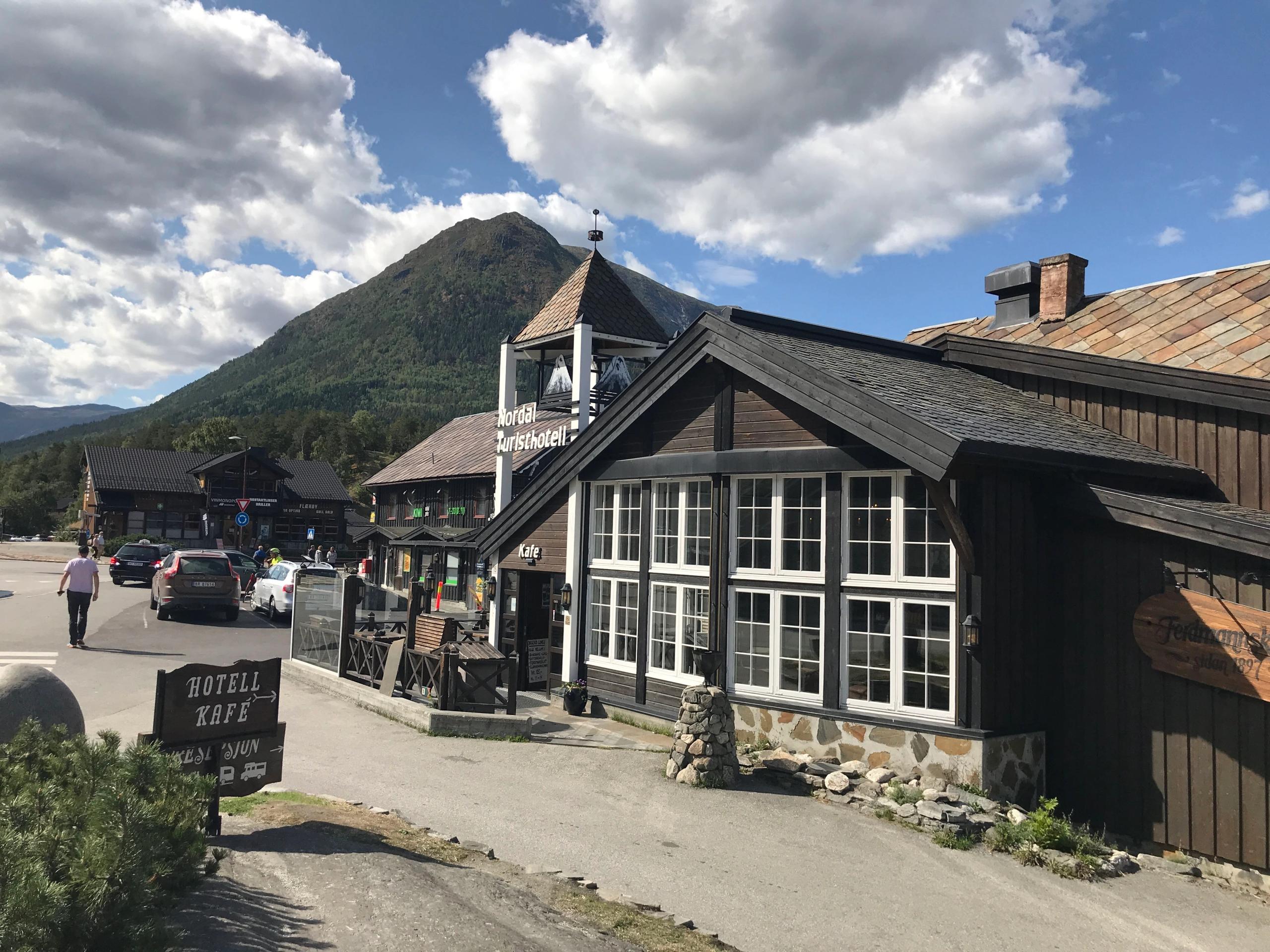 Mørk trebygning med store vindauge ved ei gate, bilar og fjell i bakgrunnen under blå himmel med skyer..