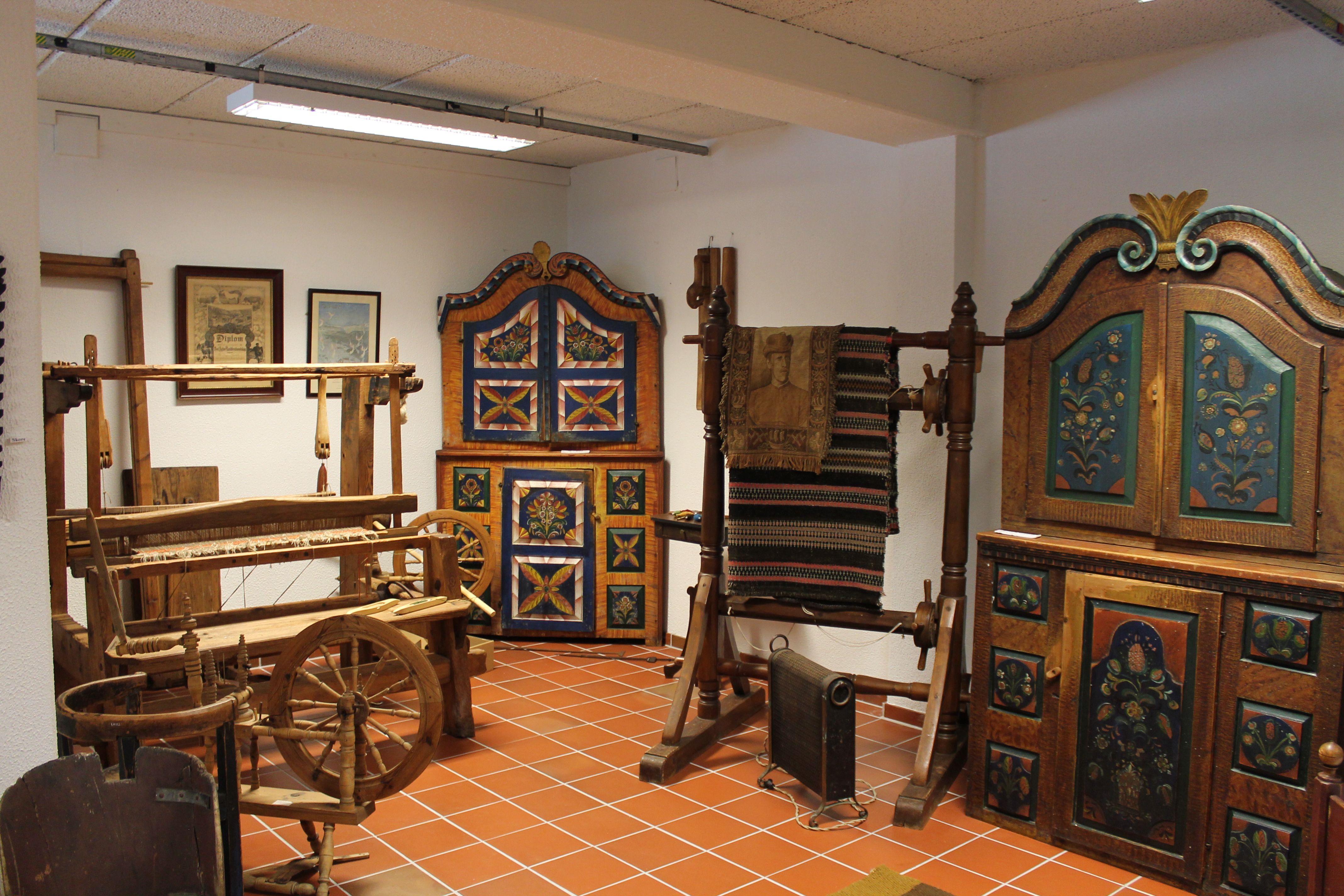 Brown cabinets with rosemaling. Weaving loom. Spinning wheel. Stand with a blanket hanging over it.