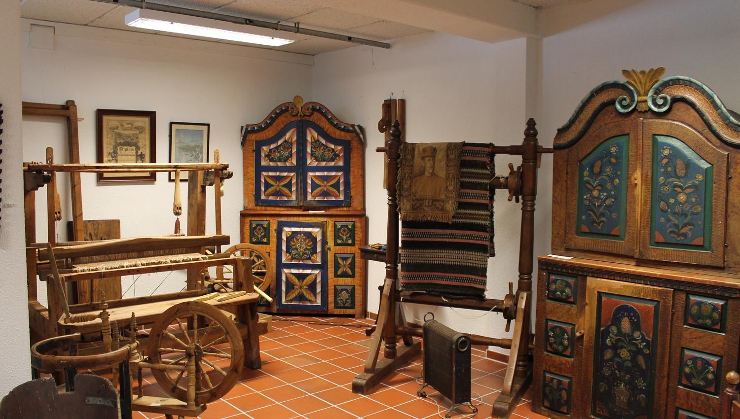 Brown cabinets with rosemaling. Weaving loom. Spinning wheel. Stand with a blanket hanging over it.