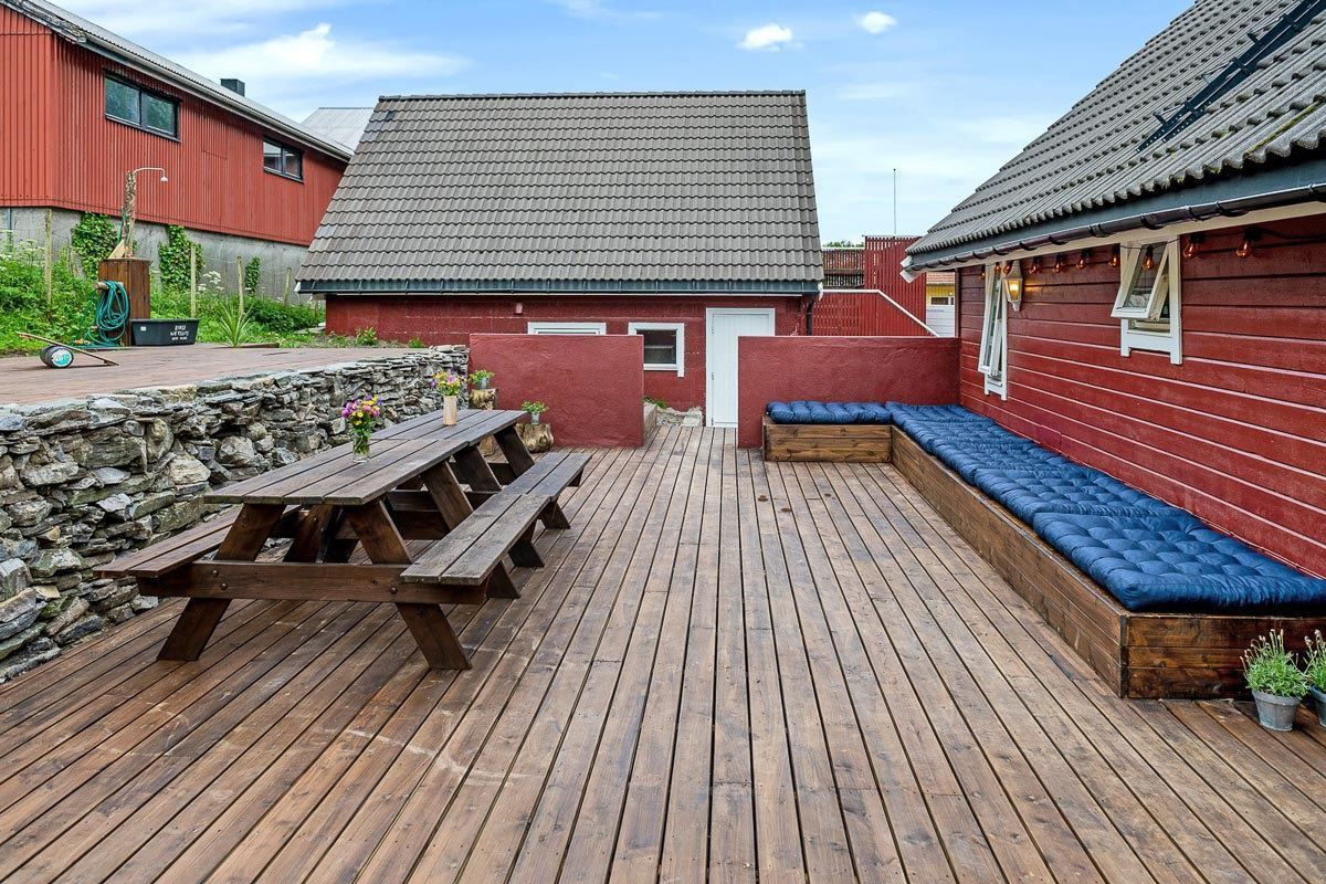 Wooden deck courtyard with a picnic table, stone wall, and long bench with blue cushions beside red buildings.