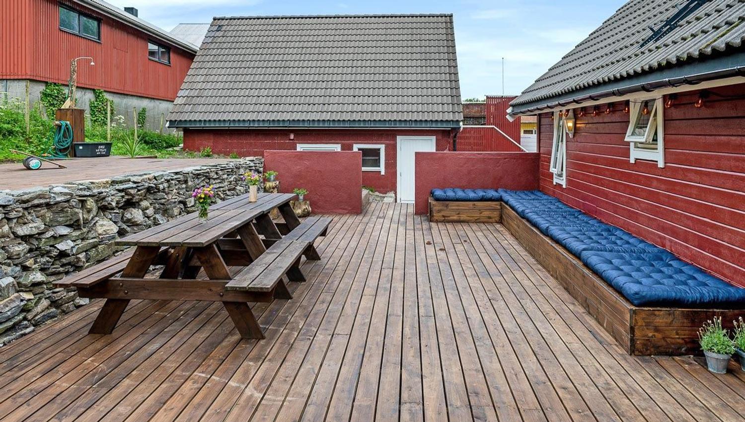 Wooden deck courtyard with a picnic table, stone wall, and long bench with blue cushions beside red buildings.