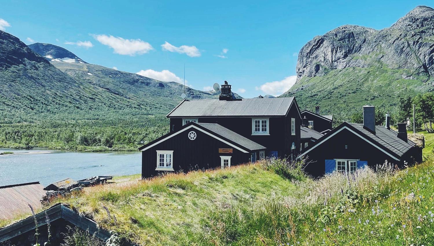 Dark wooden lodge on a grassy hillside beside a lake, with green mountains and a clear blue sky behind..