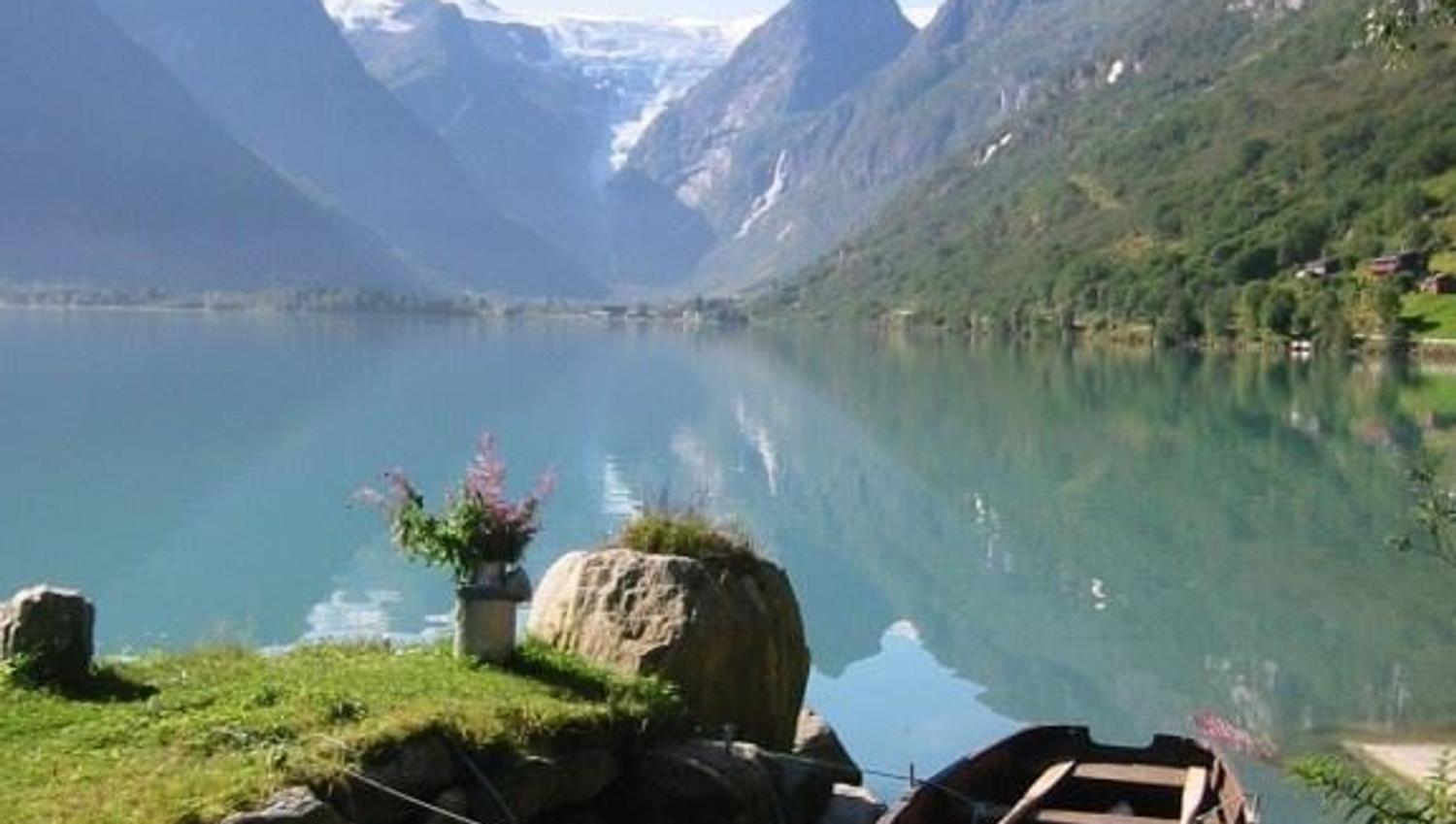 A wooden rowboat rests by a grassy shore on a calm Oldenvatnet lake, with forested mountains and distant snowfields under clear sky at Yrinset in Oldedalen, Norway..