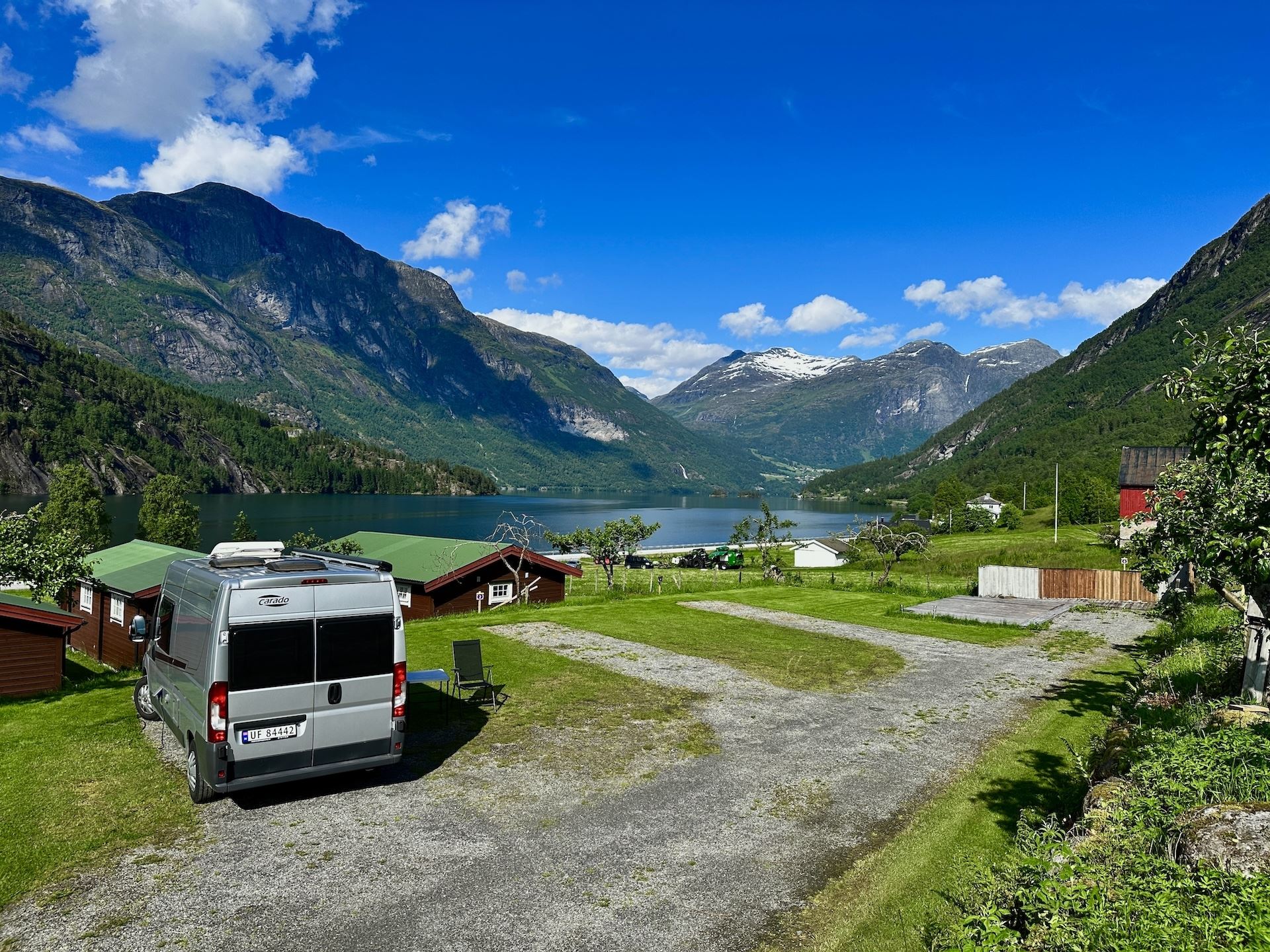 A camper van parked on grass near cabins, overlooking a calm lake and green mountains under a bright blue sky..