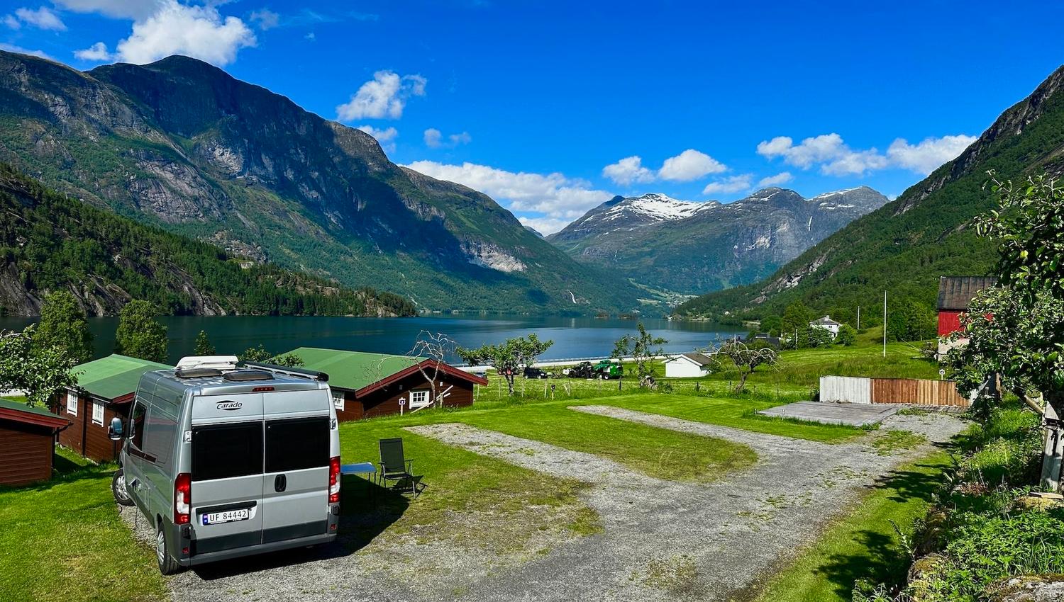 A camper van parked on grass near cabins, overlooking a calm lake and green mountains under a bright blue sky..