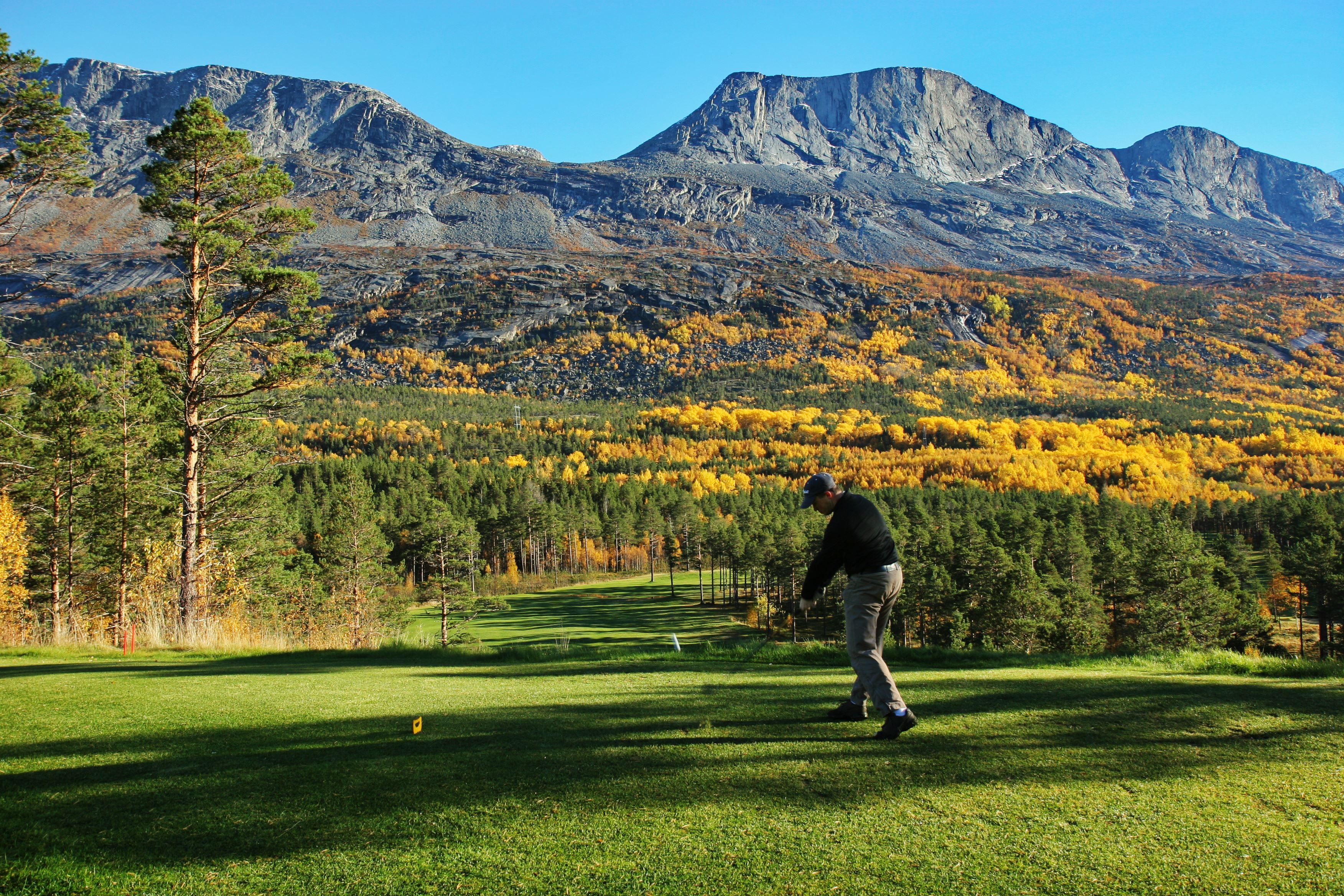 Golfer swings on a green tee box with pine trees, yellow autumn forest, and rugged mountains under clear blue sky..