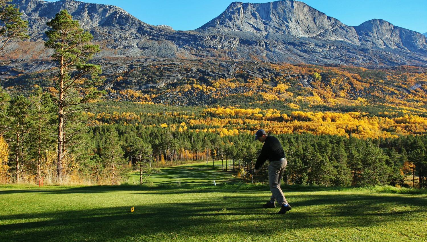 Golfer swings on a green tee box with pine trees, yellow autumn forest, and rugged mountains under clear blue sky..