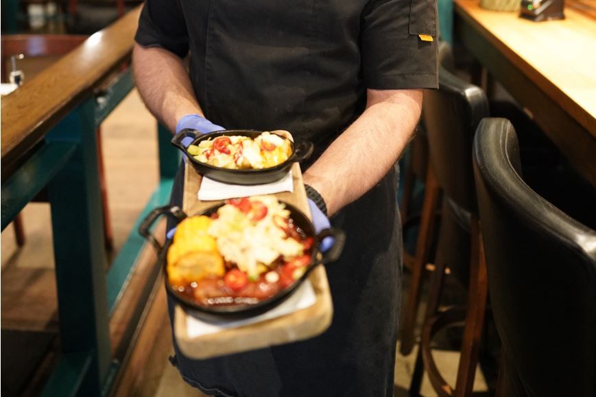 Kokk bærer to små jernpanner på et serveringsbrett, med varmrett og dessert, i en restaurant med barstoler og bord i bakgrunnen.