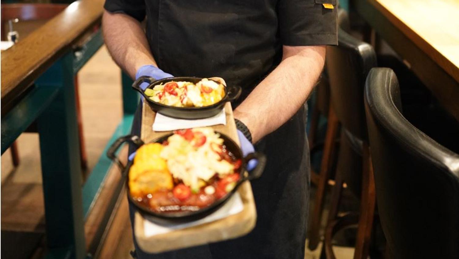 Kokk bærer to små jernpanner på et serveringsbrett, med varmrett og dessert, i en restaurant med barstoler og bord i bakgrunnen.