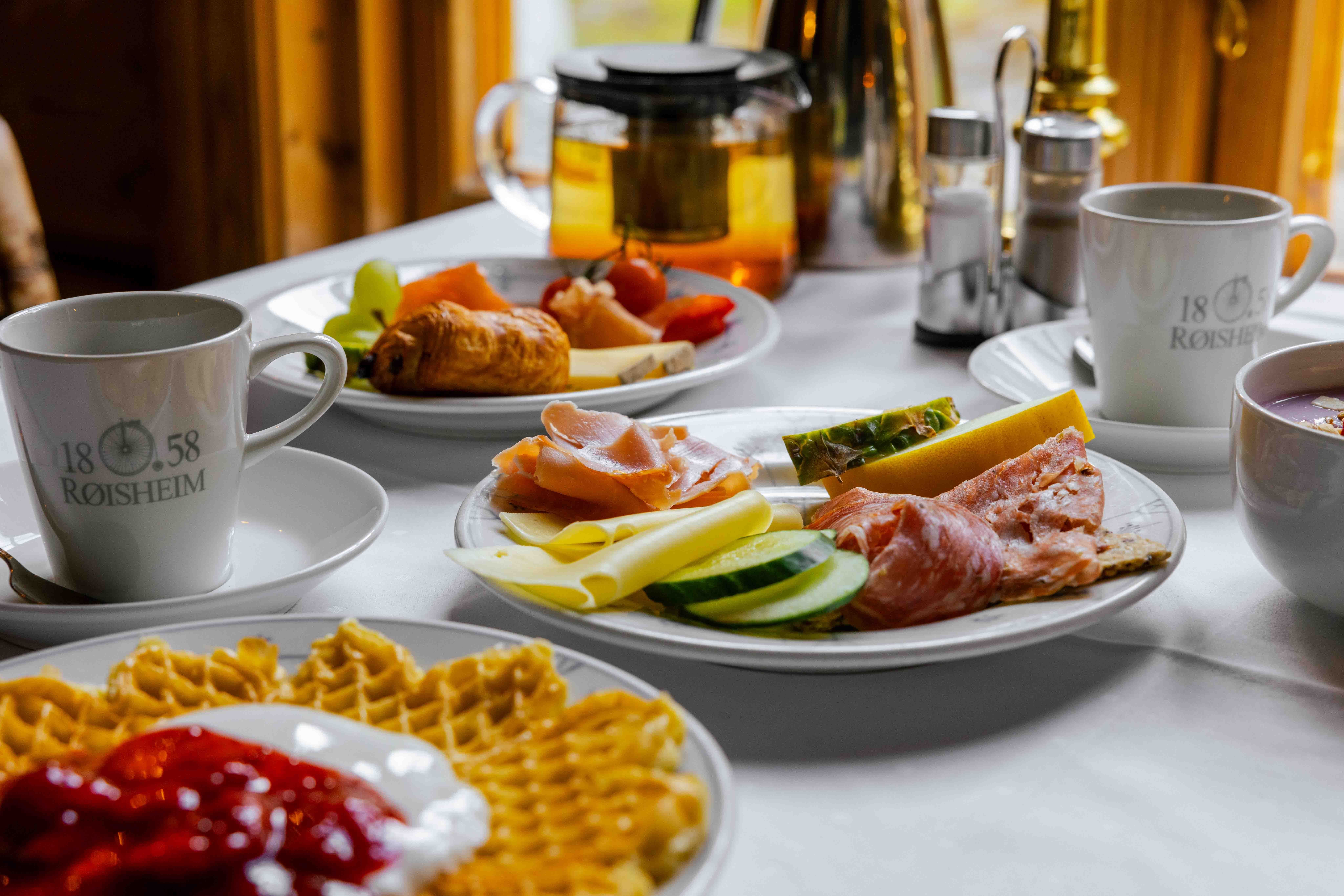 Eit frukostbord med kaffikoppar, vaflar med syltetøy, påleggsfat, tekanne og frukt på kvit duk..