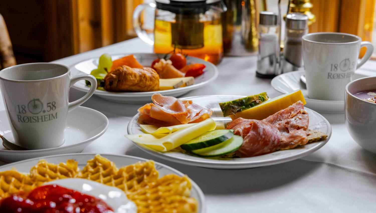 Eit frukostbord med kaffikoppar, vaflar med syltetøy, påleggsfat, tekanne og frukt på kvit duk..