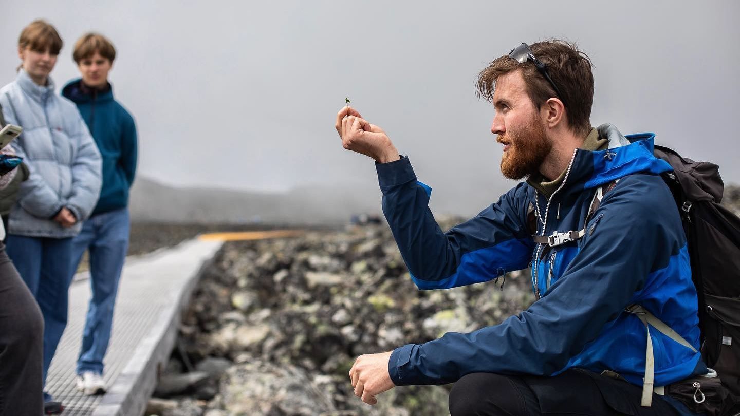 Ein person i blå jakke sit ute i steinlandskap og held ei lita plante, med andre folk i bakgrunnen..